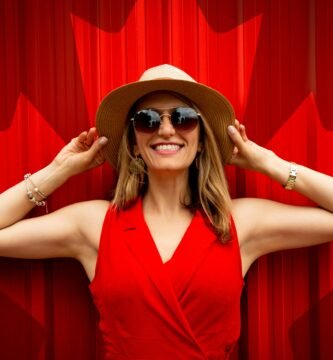 A fashionable woman in a red dress and hat smiling proudly in front of a Canadian flag backdrop.
